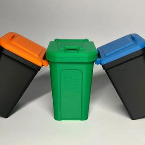 May include: Three miniature trash cans with hinged lids. One is green, the other two are black with orange and blue lids. The cans are arranged in a row, leaning slightly towards each other.
