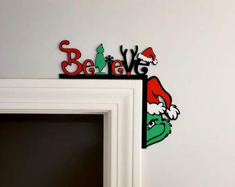 May include: A Christmas-themed corner decoration featuring the word "Believe" in red, with a green character and a red Santa hat. The decoration is black and is placed on the corner of a door frame.