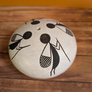 May include: A spherical, off-white decorative item featuring black stick figure designs carrying bags and tools. The object has a textured surface and small holes on top. The background is a wooden surface.