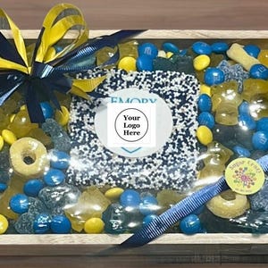 May include: A wooden box filled with an assortment of candies, including blue and yellow M&M's, gummy candies, and a frosted treat. The box is decorated with a blue and yellow bow and a ribbon. The center has a white circle with the text "Your Logo Here".
