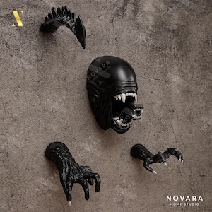 May include: Black Alien-themed wall decor set. Includes a head with an open mouth, a jaw, and two claw-like hand pieces. The pieces are mounted on a textured gray wall. The Novara Home Studio logo is visible.