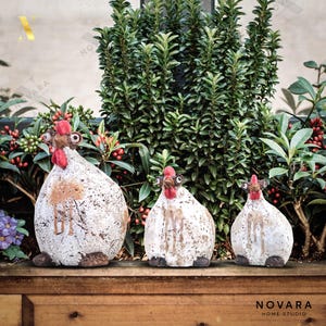 May include: Three ceramic chicken figurines with white bodies, brown accents, and red combs. The chickens have a rustic, textured appearance and are arranged in front of lush greenery. The image includes the text "NOVARA HOME STUDIO".