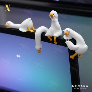 May include: Three white goose figurines with yellow feet and beaks are perched on a black surface. The geese have pink cheeks and are in various poses, including one hanging upside down. The image has the text "NOVARA HOME STUDIO".