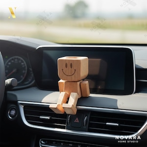 May include: A wooden figurine with a smiling face sits on a car dashboard. The figure has a block-shaped head and body with jointed arms and legs. The car's interior is black with a large display screen.