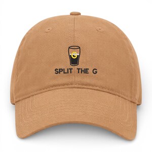 May include: A tan baseball cap with a curved brim. The cap features a black and yellow graphic of a pint glass with the text "SPLIT THE G" in black, centered below the graphic. The cap is made of a soft fabric.