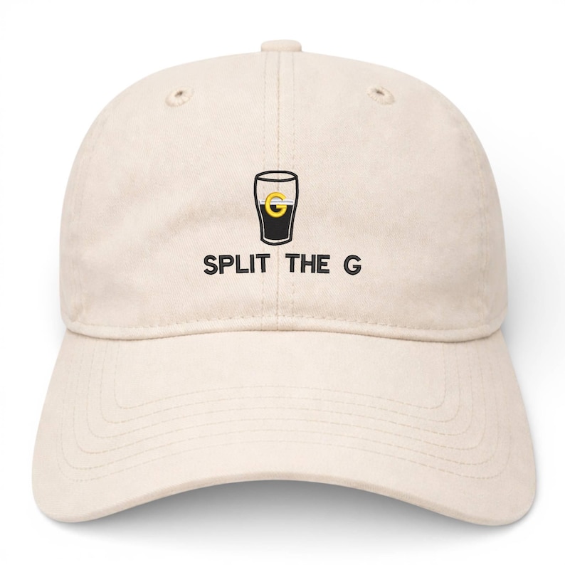 May include: A beige baseball cap with a curved brim. The cap features a black and gold graphic of a pint glass with the text "SPLIT THE G" in black, centered below the graphic. The cap has a fabric strap closure.