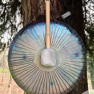 Puede incluir: Un gong grande e iridiscente con una maza de madera, colgado de un árbol. El gong tiene un tono azul y morado con líneas radiales. La maza tiene un mango de madera clara y una superficie de golpeo gris. El texto "Vento Gong VT40-0105" es visible.