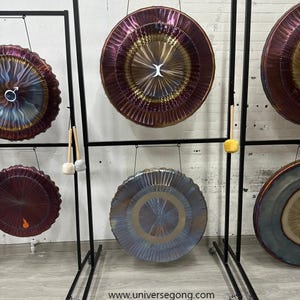 Pode incluir: Um suporte duplo de metal preto para gongos com dois gongos circulares. O gongo superior mede 85 cm e o inferior 80 cm. Texto: www.universegong.com, Double gong stand M, Top: 85cm/34" gong, Bottom: 80cm/32" gong. Inclui um martelo.