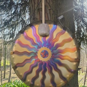 Pode incluir: Um gongo metálico circular com um padrão de raios de sol com linhas onduladas em tons de roxo, azul e laranja sobre um fundo dourado. Um martelo de madeira com cabeça cinzenta repousa no topo do gongo, que está suspenso por um cordão.