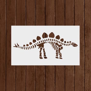 May include: A brown dinosaur skeleton stencil on a white background. The stencil depicts a Stegosaurus with its distinctive plates along its back. The image is set against a dark brown wooden background.