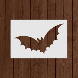 May include: A white stencil featuring a bat with outstretched wings. The bat silhouette has detailed wing edges and a small head. The stencil is on a wooden background, perfect for Halloween crafts and decorations.