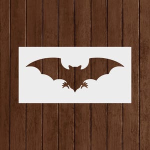 May include: A white rectangular stencil featuring a bat silhouette with outstretched wings. The bat has a detailed body and wing structure. The stencil is designed for crafting and decorative purposes, perfect for Halloween or gothic-themed projects.
