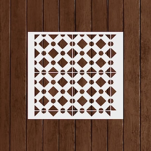 May include: A white stencil with a repeating geometric pattern of diamonds and eight-pointed stars. The stencil is square and sits against a brown wooden background. The pattern is symmetrical and creates a decorative design.
