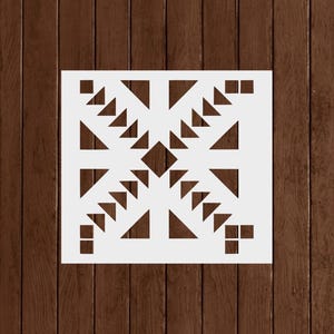 May include: A white square stencil featuring a geometric design of triangles and squares. The design is centered and symmetrical, with a diamond shape in the middle. The stencil is set against a dark brown wooden background.