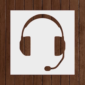 May include: A white stencil of a headset with a microphone. The headset has a curved top band and rectangular ear cups. The microphone extends from the left ear cup with a rounded end. The stencil is on a brown wooden background.