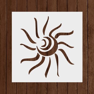 May include: A white stencil featuring a stylized sun design with curved rays and a central spiral. The sun is a warm brown color, set against a white square. The background is a dark wood grain.