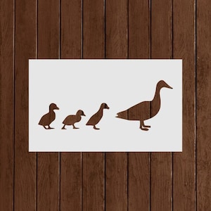 May include: A brown stencil of a mother duck followed by three ducklings on a white background. The ducks are in profile, with the mother duck facing right. The background is a wooden surface.