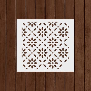 May include: A white stencil with a repeating floral pattern. The design features stylized flowers and oval shapes arranged in a grid. The cut-out pattern is brown, creating a contrast against the white background.