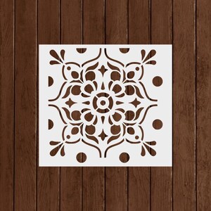 May include: A white square stencil featuring a symmetrical floral design with a central circular element and surrounding decorative patterns. The stencil includes dots and stylized leaf shapes, suitable for various craft projects.