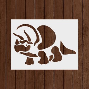 May include: A white stencil featuring a stylized Triceratops dinosaur. The design includes the dinosaur's head with horns, a frill, and a body with legs. The stencil is rectangular and set against a wooden background.
