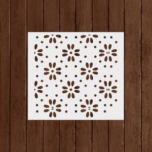 May include: A white stencil featuring a repeating floral pattern. The design includes stylized flowers and small dots, creating a decorative and symmetrical arrangement. The stencil is designed for crafts and DIY projects.