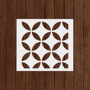 May include: A white stencil featuring a repeating pattern of overlapping, four-petal flower shapes. The stencil is square and sits on a dark brown wooden surface. The design is cut out, creating negative space.