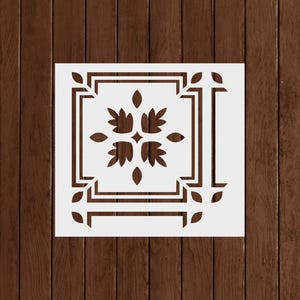 May include: A white stencil with a decorative floral and geometric design. The stencil features a central floral motif surrounded by a square frame and corner leaf accents. The background is a dark brown wood grain.