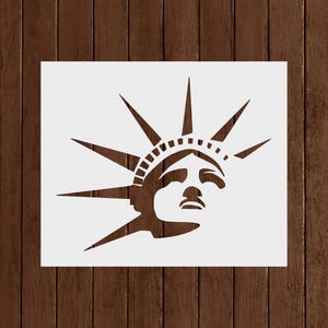 May include: A white stencil of the Statue of Liberty's face and crown. The stencil is on a white background, with a dark brown wooden surface behind it. The statue's face is in profile, with detailed lines.
