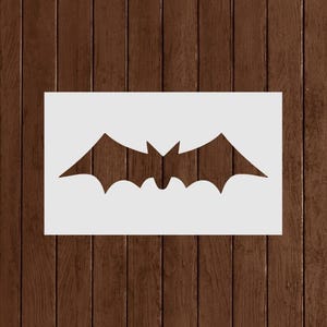 May include: A white rectangular stencil featuring a bat silhouette. The bat has pointed ears and a wide wingspan. The stencil is set against a dark brown wooden background, creating a contrast.