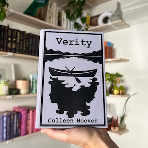 May include: A hardcover book titled "Verity" by Colleen Hoover. The cover features a black silhouette of a boat on water, with a landscape and two faces. The book is held in front of a white wall with shelves of books and plants.