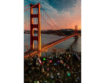 May include: A view of the Golden Gate Bridge in San Francisco, California. The bridge is red and orange and is covered in a layer of fog. The bridge is surrounded by water and there are many locks attached to a fence in the foreground.