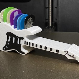 May include: A white guitar pick holder with a black pickguard design, holding several colorful guitar picks. The word "Fender" is printed on the neck of the holder. A practical and decorative item for guitar players.