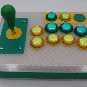 May include: A custom arcade controller with a green joystick and yellow accents. It features several colorful buttons: yellow, green, and teal. The controller is housed in a clear, textured case with green trim.