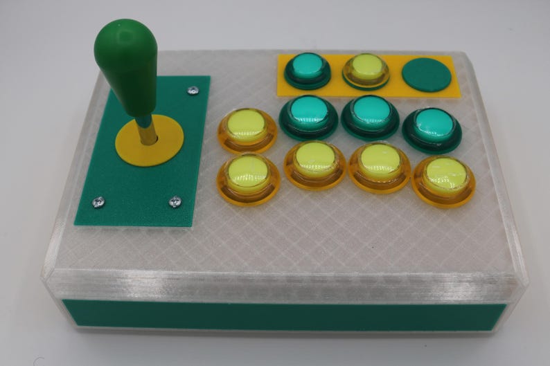 May include: A custom arcade controller with a green joystick and yellow accents. It features several colorful buttons: yellow, green, and teal. The controller is housed in a clear, textured case with green trim.