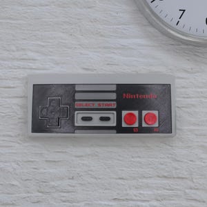 May include: A gray and black Nintendo controller with red buttons and the word "Nintendo" in red. The controller has a directional pad, select and start buttons, and two red action buttons. The controller is mounted on a white brick wall.