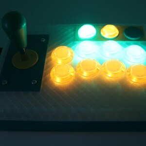 May include: A custom-built arcade controller with a green joystick and yellow accents. It features eight illuminated buttons in yellow and white, and a green button. The controller is designed for gaming.