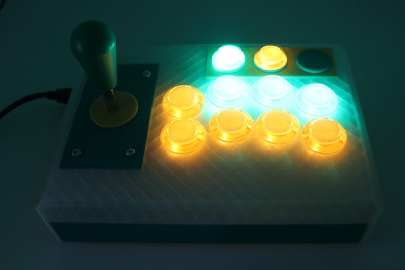 May include: A custom-built arcade controller with a green joystick and yellow accents. It features eight illuminated buttons in yellow and white, and a green button. The controller is designed for gaming.