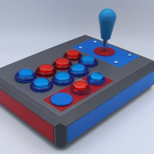 May include: A custom arcade game controller with a blue joystick and red and blue buttons. The controller has a gray body with red and blue accents. The buttons are arranged in a classic layout.