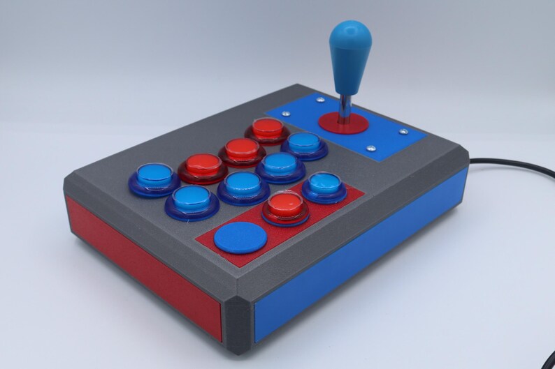 May include: A custom arcade game controller with a blue joystick and red and blue buttons. The controller has a gray body with red and blue accents. The buttons are arranged in a classic layout.