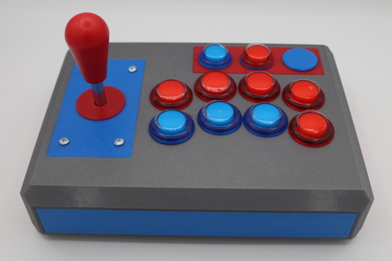 May include: A gray arcade game controller with a red joystick and buttons. The controller has a blue base and accents, with red and blue buttons arranged in a classic layout. The joystick is on the left side.