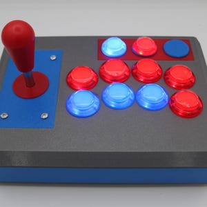 May include: A gray arcade game controller with a red joystick and several illuminated buttons. The buttons are red and blue, and the controller has a blue accent on the front and sides.