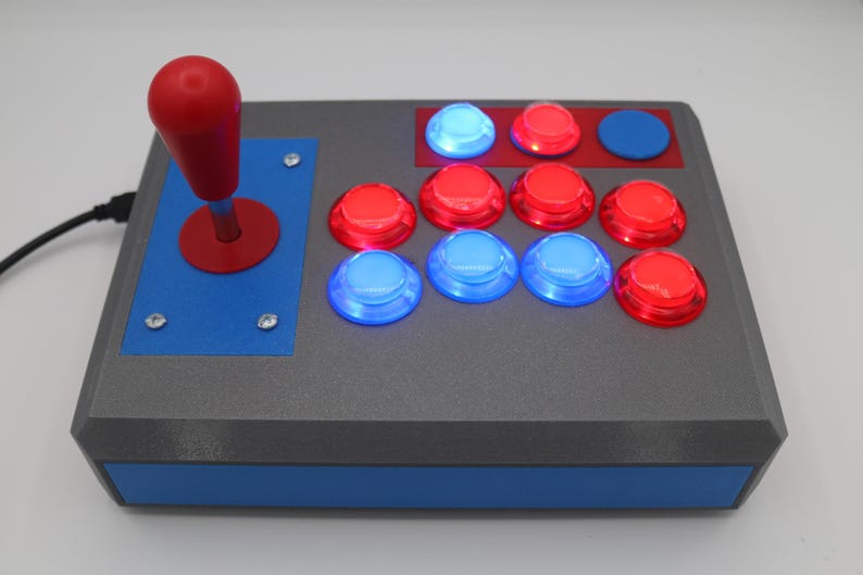 May include: A gray arcade game controller with a red joystick and several illuminated buttons. The buttons are red and blue, and the controller has a blue accent on the front and sides.
