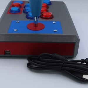 May include: A gray arcade game controller with red and blue accents. The controller has a blue joystick and several red and blue buttons. A black USB cable is connected to the controller.