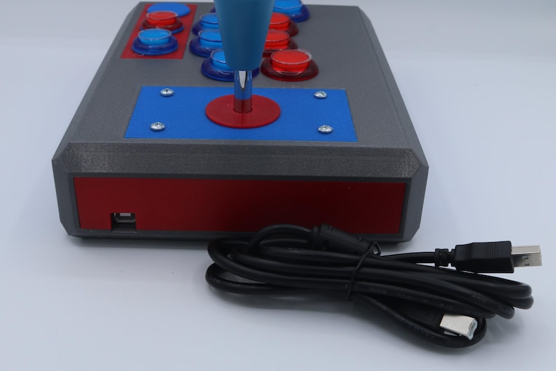 May include: A gray arcade game controller with red and blue accents. The controller has a blue joystick and several red and blue buttons. A black USB cable is connected to the controller.
