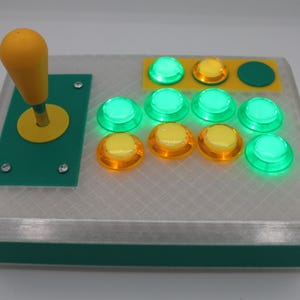 May include: A custom arcade controller with a yellow joystick and buttons. The buttons are a mix of green and yellow, and the controller has a green and white case. The joystick is on the left side of the controller.