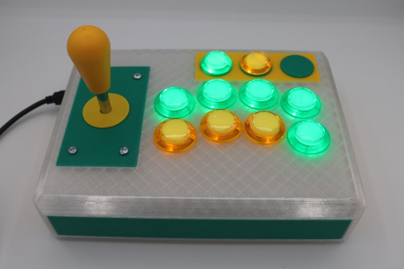 May include: A custom arcade controller with a yellow joystick and buttons. The buttons are a mix of green and yellow, and the controller has a green and white case. The joystick is on the left side of the controller.