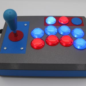 May include: A gray arcade game controller with a blue joystick and red and blue buttons. The controller has a blue base and a black cable. The buttons are arranged in two rows, with the top row having three buttons and the bottom row having six buttons.