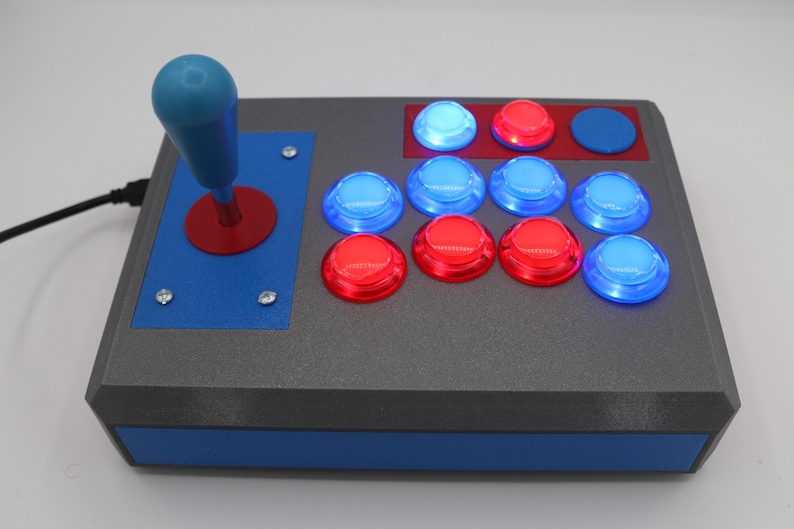 May include: A gray arcade game controller with a blue joystick and red and blue buttons. The controller has a blue base and a black cable. The buttons are arranged in two rows, with the top row having three buttons and the bottom row having six buttons.