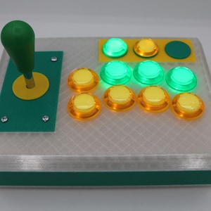 May include: A custom-built arcade game controller with a green joystick and buttons. The controller has a clear, textured top and a green base. The buttons are a mix of yellow and green, some of which are illuminated.