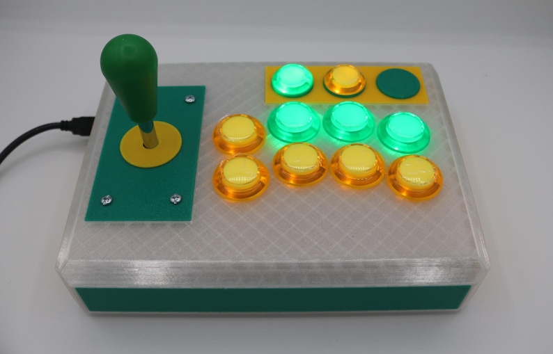 May include: A custom-built arcade game controller with a green joystick and buttons. The controller has a clear, textured top and a green base. The buttons are a mix of yellow and green, some of which are illuminated.
